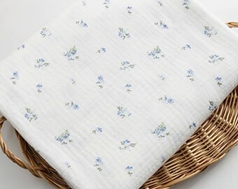 Op de afbeelding: Witte stof met een klein blauw bloemenpatroon. De stof is gevouwen en op een gevlochten mand gelegd.
