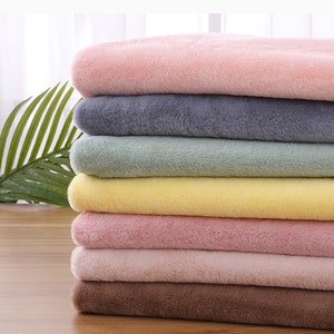 Może przedstawiać: Stosik siedmiu próbek pluszowej tkaniny w różnych kolorach, w tym różowym, szarym, zielonym, żółtym, brązowym i jasnoróżowym. Próbki są złożone i ułożone na drewnianej powierzchni.