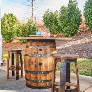 May include: A wooden barrel bar table with a round top and two matching barrel stools. The table and stools are made from reclaimed wood and have a rustic finish. The table is set with a bottle of liquor, two glasses, and a small wooden tray.