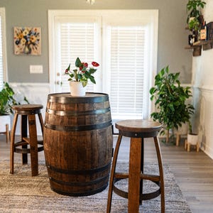 May include: A rustic, wooden barrel table with metal bands, topped with a white pot of red flowers. Two wooden stools with round seats and angled legs flank the table. The scene is set on a textured rug.