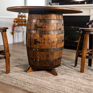 May include: A rustic bar table and two matching stools. The table is made from a repurposed wooden barrel with metal bands and a round tabletop. The stools have wooden legs and circular seats. The furniture is brown and sits on a neutral-colored rug.