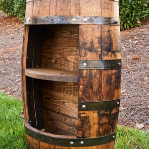 May include: A wooden barrel with metal bands and two shelves. The barrel is brown and has a dark stain. The shelves are made of wood and are stained a darker brown. The barrel is sitting on a green grassy surface.