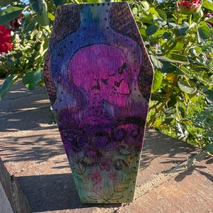 Puede incluir: Una caja de madera con forma de ataúd con un diseño pintado a mano. El ataúd presenta un cráneo y elementos florales en tonos morados, azules y verdes. La obra de arte está sobre un fondo de vetas de madera natural.