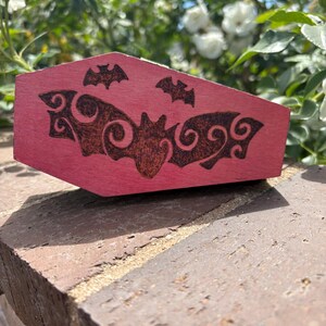 Puede incluir: Una caja de madera rosa con forma de ataúd con un diseño de murciélago. La caja tiene un diseño de murciélago marrón oscuro en la tapa, con dos murciélagos más pequeños encima del más grande. La caja está sobre una superficie de ladrillo.