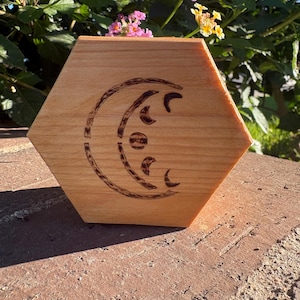 Puede incluir: Caja hexagonal de madera clara con un diseño de luna creciente quemado. El diseño presenta un círculo central y varias formas de media luna. La caja está sobre una superficie de ladrillo.