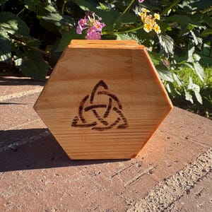 Puede incluir: Una caja hexagonal de madera con un diseño de nudo celta quemado en la madera. La caja está hecha de madera de color claro y tiene un acabado natural. El fondo incluye follaje verde y flores rosas y amarillas.