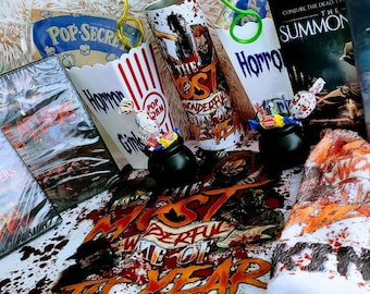 May include: A collection of horror-themed items including two DVDs titled Dark Whispers and The Summoned, a popcorn bucket, a tumbler with horror graphics, two small black cauldrons filled with wrapped candies, and a white blanket with red blood splatter.