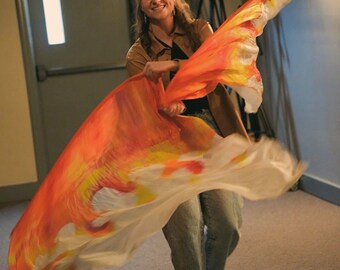May include: A woman is holding a large, flowing silk flag. The flag features a vibrant design with shades of orange, yellow, and white, resembling flames. The flag is in motion, creating a dynamic and energetic visual.