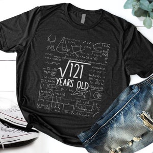 May include: A dark grey t-shirt with a white graphic design of mathematical equations and the text "√121 YEARS OLD".