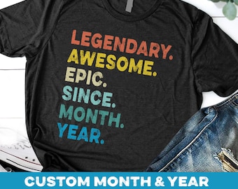 May include: A black t-shirt with a vintage-style graphic that reads "Legendary. Awesome. Epic. Since. Month. Year."  The text is in rainbow colors.  The shirt is laid flat on a white surface with a pair of jeans and a white sneaker.