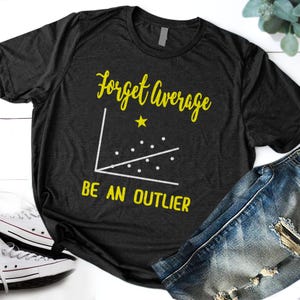May include: A charcoal gray short-sleeve t-shirt featuring a yellow graphic print with the text "Forget Average" and "BE AN OUTLIER" surrounding a white scatter plot graph with a single yellow star positioned above the data points.