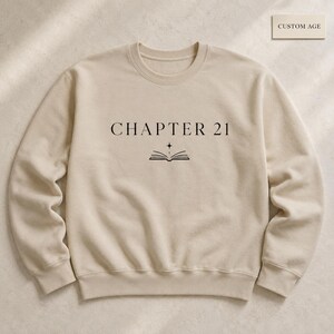 May include: A beige crewneck sweatshirt featuring the black text "CHAPTER 21" centered on the chest, accompanied by a small graphic of an open book with a four-pointed star above it. The garment has long sleeves and a ribbed collar, cuffs, and hem.