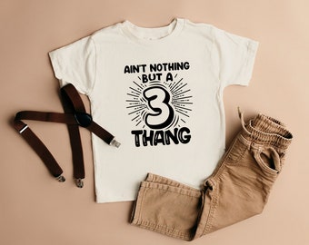 May include: White t-shirt with black text that reads "Ain't nothing but a 3 thang". The number 3 is stylized with a sunburst behind it. The shirt is laid flat on a light brown surface with brown suspenders and brown pants.