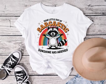 Può includere: Maglietta bianca con un procione dei cartoni animati, un arcobaleno e la scritta "YOUR LITTLE RAY OF SARCASTIC SUNSHINE HAS ARRIVED". L'immagine include scarpe da ginnastica nere e un cappello beige.