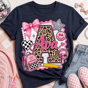 May include: A navy blue t-shirt featuring a graphic print of a large letter A with a leopard print pattern, the name Ava in pink script, a pink bow, a pink backpack, a yellow pencil, a checkered heart, a glittery star, and a disco ball.