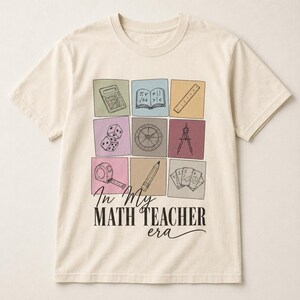 Peut inclure: Un t-shirt à manches courtes couleur crème avec une grille de neuf carrés colorés contenant des dessins au trait d'une calculatrice, d'un livre, d'une règle, de dés, d'un compas, d'un rapporteur, d'un mètre ruban, d'un crayon et de cartes.