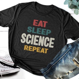 Könnte beinhalten: Dunkelgraues T-Shirt mit dem Aufdruck "EAT SLEEP SCIENCE REPEAT" in Rot, Türkis, Weiß und Gelb. Das Shirt wird mit blauen Jeans und weißen Turnschuhen kombiniert.