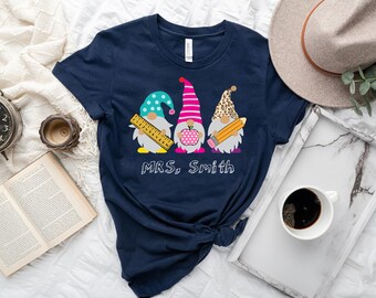 May include: A navy blue t-shirt featuring a design of three gnomes wearing school supplies. The gnomes are holding a ruler, an apple, and a pencil. The text "MRS. Smith" is printed below the gnomes.