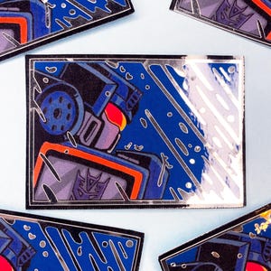 Puede incluir: Un pin rectangular de esmalte que muestra un robot azul con ojos rojos y un logotipo Decepticon morado en el pecho. El diseño incluye detalles metálicos plateados y líneas diagonales sobre un fondo azul oscuro, capturando una ilustración gráfica estilizada de la figura robótica.
