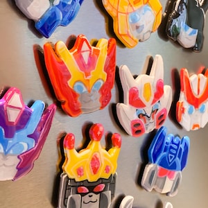 May include: A selection of hand-painted robot head magnets in various colours. The magnets display diverse designs with bright hues such as blue, red, yellow, and white. Each magnet showcases a unique robot face design, with some featuring crowns.