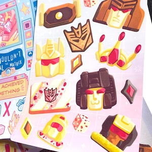 May include: Sheet of stickers featuring various cartoon character designs in shades of yellow, brown, and red. The stickers include character faces, symbols, and geometric shapes. The background is a light pink grid pattern.