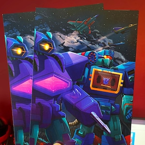 May include: A purple and blue robot with a cassette tape on its chest stands next to another purple robot with glowing eyes. Both robots are standing on a red and orange surface with a dark blue background.