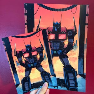Puede incluir: Dos impresiones de Optimus Prime, un robot negro y rojo, se alzan sobre un fondo naranja y azul. El robot está en una pose dinámica, con un paisaje urbano visible a lo lejos. Las impresiones se sostienen contra una pared roja.