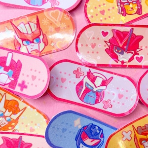 May include: A collection of colorful adhesive bandages featuring cartoon robot character designs. The bandages have various background colors, including pink, yellow, and blue, with heart and cross motifs. The robot characters are in shades of pink, red, orange, and blue.