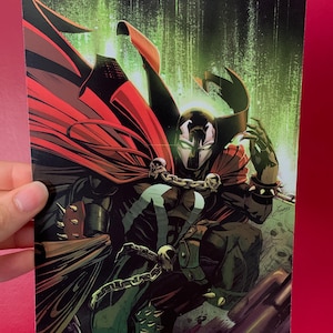 Puede incluir: Ilustración de cómic de Spawn, un superhéroe, en una pose dinámica. El personaje está vestido con un traje negro con una capa roja, cadenas y púas. El fondo es una mezcla de verde y negro, con una máscara con forma de calavera.
