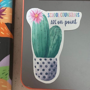 May include: A sticker with a watercolor illustration of two cacti in a pot with black polka dots. The cacti are green and have pink flowers. The text on the sticker reads "School Counselors Are on Point".