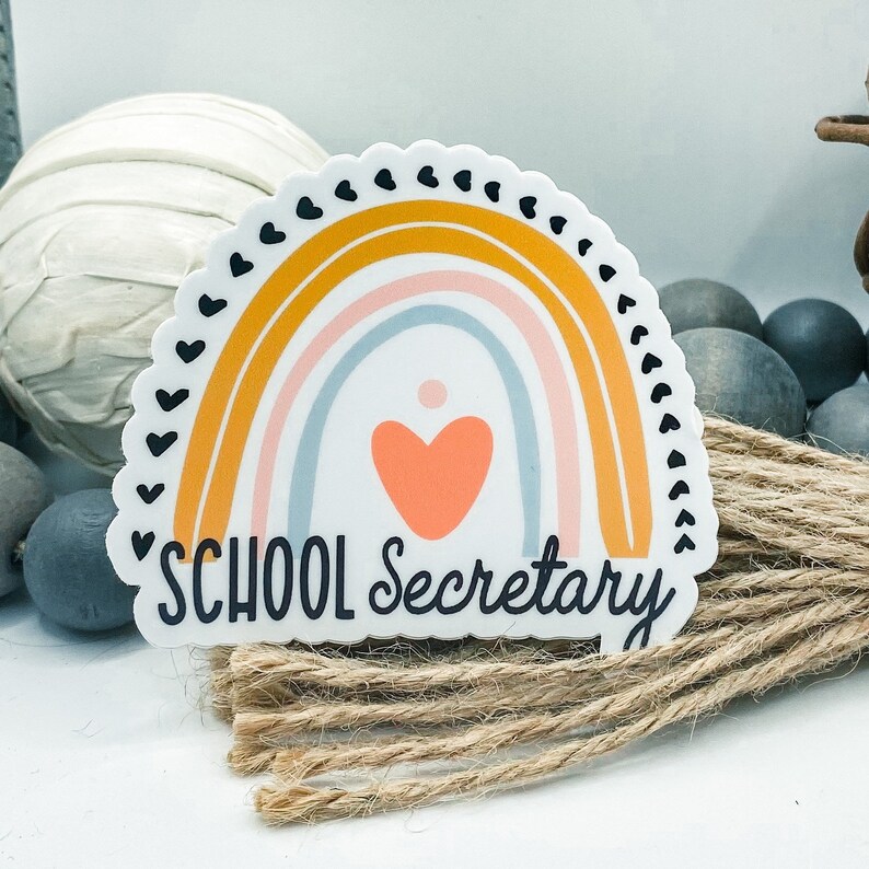 May include: A white sticker with a rainbow design in shades of yellow, pink, and blue. The rainbow has a heart in the center and the text "SCHOOL Secretary" at the bottom.