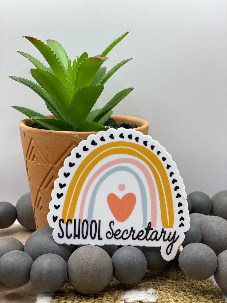 May include: A white sticker with a rainbow design and the text "SCHOOL Secretary" in black. The rainbow has a pink, blue, and yellow color scheme with a small orange heart in the center.