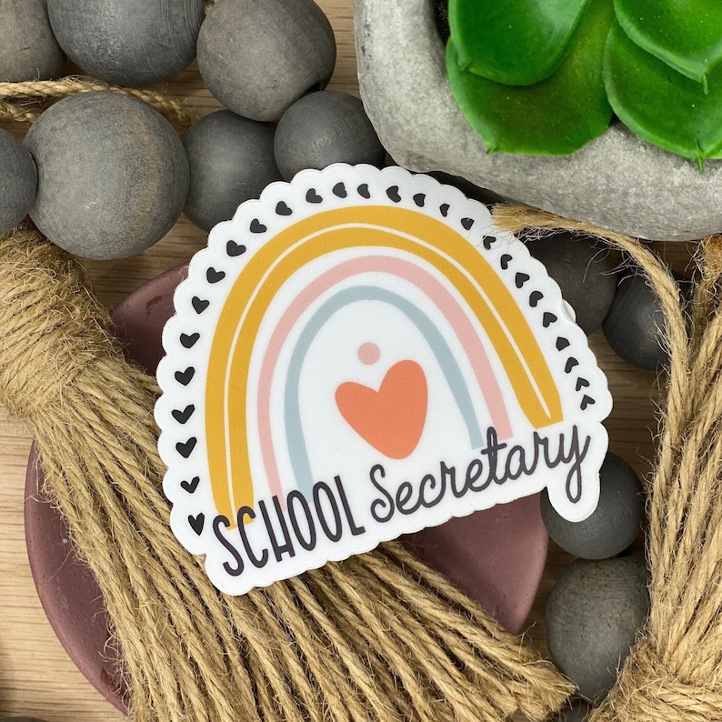 May include: A sticker with a rainbow design in yellow, pink, and blue with a heart in the center. The text "SCHOOL SECRETARY" is written below the rainbow.