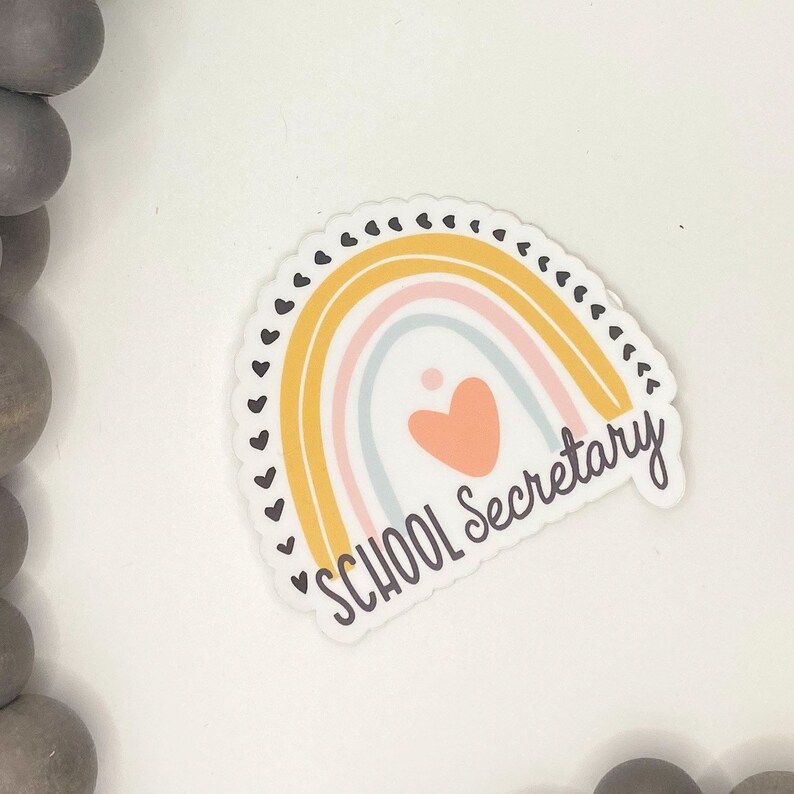 May include: A sticker with a rainbow design and the text "SCHOOL Secretary" in black lettering. The rainbow is made up of yellow, pink, and blue stripes with a small pink heart in the center.