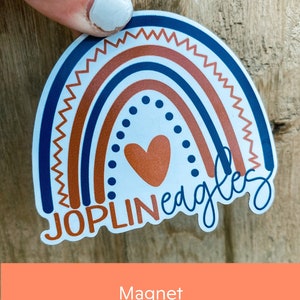 May include: A round magnet with a rainbow design in shades of blue, orange, and white. The text "JOPLINeagles" is written in a stylized font with a heart in the center of the rainbow.