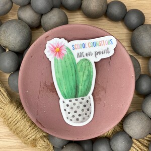 May include: A sticker with a cactus and the text "School Counselors Are on Point" in rainbow colors. The cactus is green with a pink flower and is in a gray pot with white polka dots.