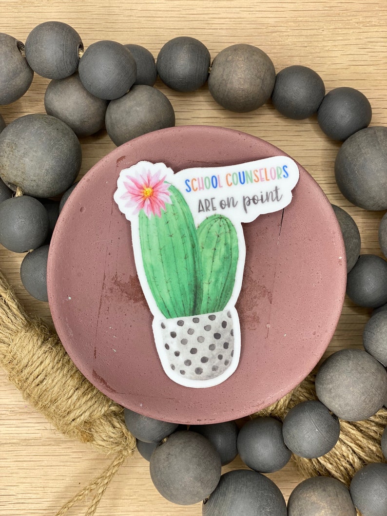 May include: A sticker with a cactus and the text "School Counselors Are on Point" in rainbow colors. The cactus is green with a pink flower and is in a gray pot with white polka dots.