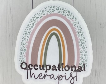 May include: A sticker with a rainbow design and the words "Occupational therapist". The rainbow features shades of pink, brown, yellow, and green, with a leopard print border. The sticker is white with a rounded edge.