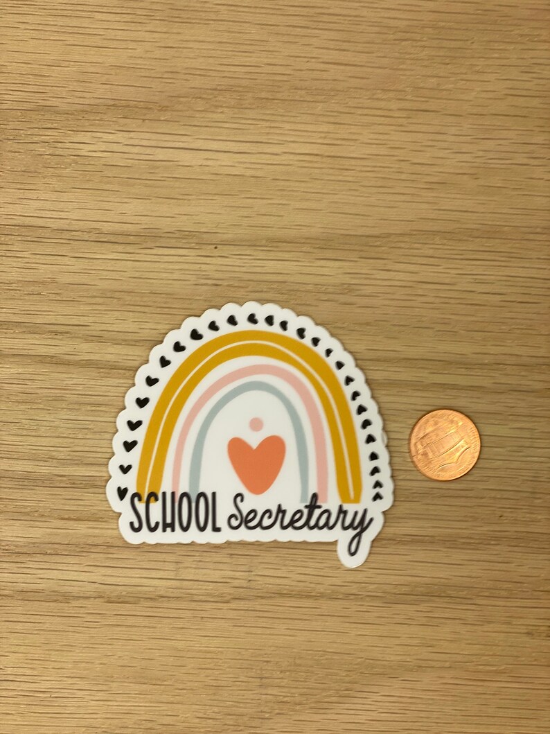 May include: A white sticker with a rainbow design and the text "SCHOOL Secretary". The rainbow is made of yellow, orange, pink, and blue stripes. The sticker is on a wood background.