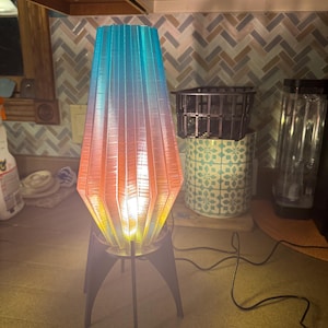 May include: A modern, geometric lamp with a pleated, ombre shade in shades of blue, orange, and yellow. The lamp is on a black metal tripod stand.