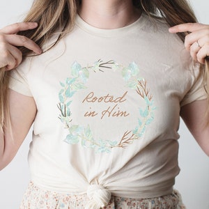 May include: A cream-colored t-shirt with a floral wreath design and the text "Rooted in Him" in brown lettering.
