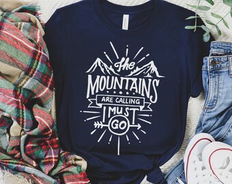 May include: A navy blue t-shirt with a white graphic that says "The Mountains Are Calling I Must Go". The graphic features a mountain range, an arrow, and a sunburst. The t-shirt is laid out on a white surface with a plaid scarf, blue jeans, and white sneakers.