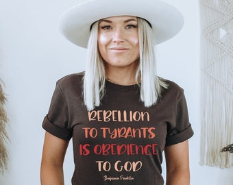 May include: A woman wearing a brown t-shirt with the text "Rebellion to Tyrants is Obedience to God" and a white hat. The text is in a red and orange gradient.
