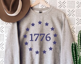 May include: A gray sweatshirt with a blue circle of stars around the number 1776. The sweatshirt is hanging on a wooden hanger against a white background with a black geometric pattern.