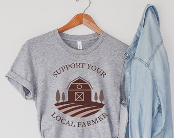 May include: A gray t-shirt with the text "Support Your Local Farmer" and a graphic of a barn with trees. The shirt is hanging on a wooden hanger with a denim jacket draped over it. A pair of light blue denim shorts are also pictured.