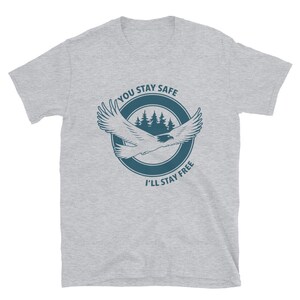 K&ouml;nnte beinhalten: Ein graues T-Shirt mit einem blauen Kreis-Motiv, das einen fliegenden Adler, Kiefern und den Text "You Stay Safe I'll Stay Free" zeigt.