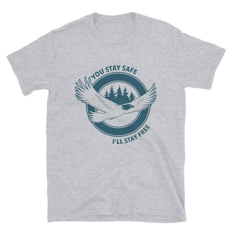 K&ouml;nnte beinhalten: Ein graues T-Shirt mit einem blauen Kreis-Motiv, das einen fliegenden Adler, Kiefern und den Text "You Stay Safe I'll Stay Free" zeigt.
