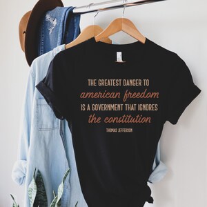 May include: Black t-shirt with a quote by Thomas Jefferson in brown text: "THE GREATEST DANGER TO american freedom IS A GOVERNMENT THAT IGNORES the constitution"