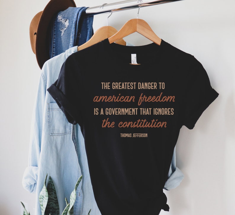 May include: Black t-shirt with a quote by Thomas Jefferson in brown text: "THE GREATEST DANGER TO american freedom IS A GOVERNMENT THAT IGNORES the constitution"