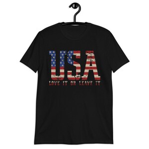 Pu&ograve; includere: Maglietta nera con un design di bandiera americana usurato che recita "USA LOVE IT OR LEAVE IT".